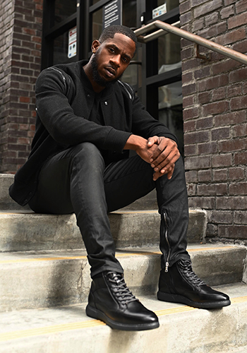 Image of social media influencer Chidi Ezemma sitting on  steps outside while wearing the Flair Moc Toe Lace Up Boot in Black.
